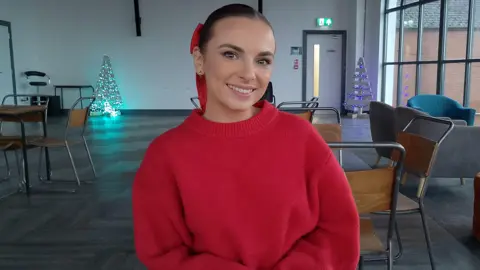 BBC Amber Harrison is wearing a red jumper and has a red bow in her brown hair. She's sitting in a large room, with some Christmas tree decorations in the background. 