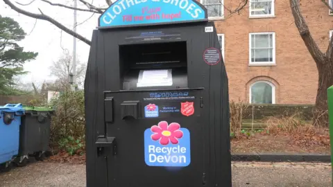 A black bin that says "clothing & shoes". Behind is to the left are two bins, one blue and one black, to the right is a tree and a brown brick building. 
