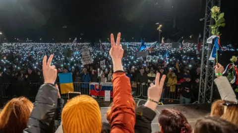 An evening protest in EPA Bratislava, where several protesters with their backs to the camera gave the victory salute and one held a rose. They encounter a large group of protesters behind barriers holding banners and Slovakian and Ukrainian flags