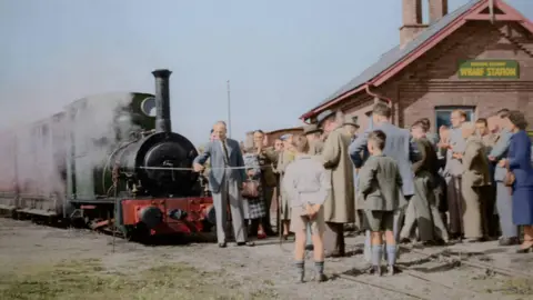 Talyllyn Railway collection An old picture from 1951 of a small green steam locomotive with a rake of carriages standing on some track in a yard. A ribbon is stretched in front of it, and a man in a suit is preparing to cut the ribbon while a sizeable crowd watches. All are dressed in styles typical of the period.