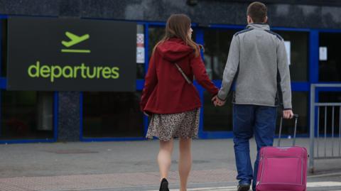 A couple hold hands as they walk towards the entrance to Belfast International Airport