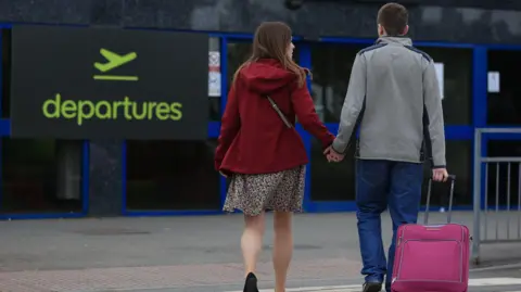 PA A couple hold hands as they walk towards the entrance to Belfast International Airport