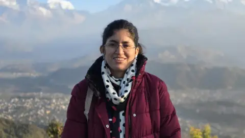 Usama Noor Safkat Umama Fatema wears a maroon coat and polka dot scarves and smiles at the camera at a sightseeing spot, in the background is a mountain range
