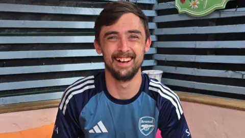 An image of smiling Adam McClelland with a birthday cake in front of him, in celebration of his 26th birthday. He is holding a glass with a drink. He is sitting in what appears to be an outdoor area.