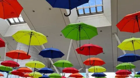 Hester Grainger A number of colourful open umbrellas hanging from the roof of a shopping, seen from below. They are in bold colours: red, blue, yellow and green with black handles. 