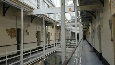 The inside of one of the prison wings. It's a mezzanine with rows of doors on both sides and metal pillars down the middle. In some areas, the white paint and plaster is coming off the walls and exposing the brick.