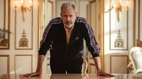 Justin Downing/Netflix Jeff Goldblum leaning forward with his hands on a table in an opulent-looking room with candlebras on the wall behind him