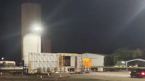 A large wooden structure can be seen lit by large floodlights on an industrial dock, including a large concrete tower. 