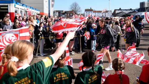 Gloucester-Hartpury players arrive at Kingsholm ahead of their match with Bristol Bears women. It is a sunny, clear day and hundreds of fans are there to greet them, some waving flags.