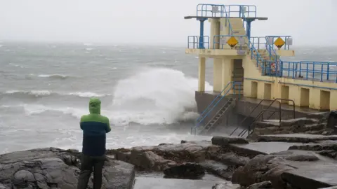 PA Media A person wearing a green and blue coat stands with their back to the camera watching waves crash over a bathing spot on the coast.