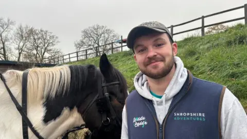 BBC/ Alex James A bearded man stands to the right of the frame wearing a blue body warmer with a logo over a grey hoodie. He is wearing a hat. Next to him there is a white and black horse. 