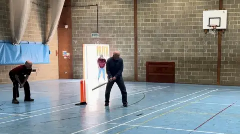 Sandy Cricket Club Indoor sports hall with marked court lines where several adults are playing walking cricket. One person is holding a bat near bright orange stumps and others are fielding. A basketball hoop is mounted on a brick wall in the background and an open doorway is letting in daylight.
