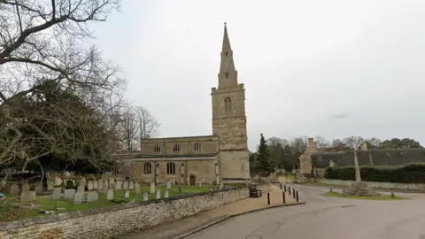 Google A google streetview shot of a church with a tower and a graveyard in front of it. A road runs alongside the church.
