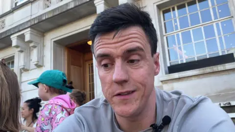 Paul Biggin, a man with dark hair and wearing a grey top, he is sitting in front of Hackney Town Hall