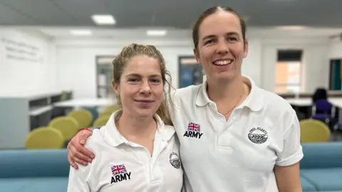 Emma Gibb and Imy O'Brien stand in the centre of the frame. They are both wearing white polo shirts that feature the Union Flag and the word "ARMY" in block capitals. They stand in a classroom with empty chairs visible behind them. Imy who is the taller of the two has her arm around Emma. 