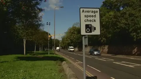 A average speed check sign alongside a road with cameras on a yellow pole further down the road