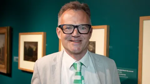 Qays Najm/BBC A man smiles at the camera at an art exhibition. He has short grey hair and is wearing a light grey suit, glasses, white shirt and a green and white tie. Pieces of art are on the wall behind him.