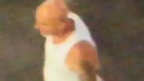 PSNI The image is of CCTV footage of a man allegedly involved in disorder in Ballymena. He is wearing a white vest top and has several tattoos on his left arm. His head is completely shaved.