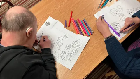 Vikki Irwin/BBC The picture is from above over a wooden table, where two people are seated. Each person is colouring in large printed drawings on white paper. The person on the left is using a dark-coloured marker and has a hearing aid visible. The person on the right is using a purple felt-tip pen. Several other felt-tip pens are scattered across the table. 
