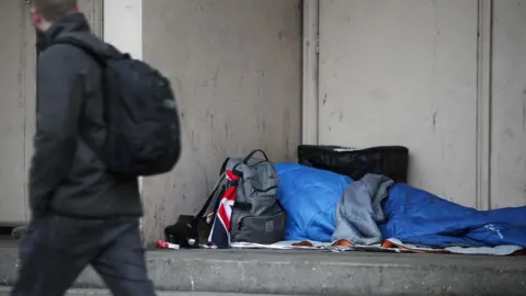 A man walks past a sleeping bag and a person's belongings on the street.