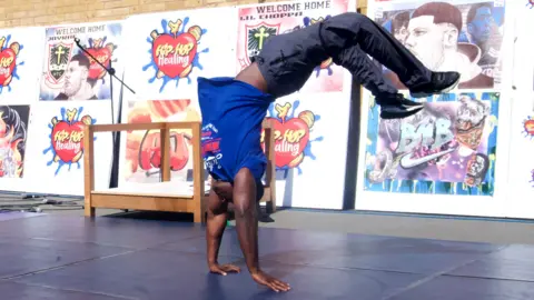 A man in a blue t-shirt and grey trousers performing a back flip on a blue mat