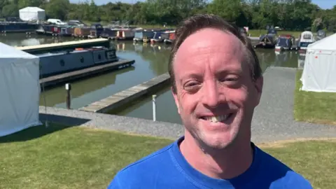 Annabel Amos/BBC A man in a blue T-shirt smiles at the camera. Behind him is a marina with lots of narrowboats docked.