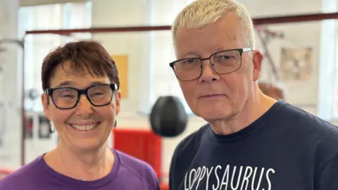 Simon Thake/BBC A woman with short dark hair and black-rimmed glasses smiles at the camera next to an unsmiling man with white hair, also wearing glasses.