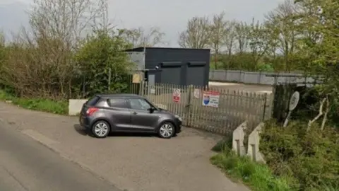 Google Grey prefab buildings with roller shutters in a plot alongside a rural road. The site is surrounded by trees and there are metal gates across the entrance, which have security company warning notices on them. A small grey car is parked at the entrance.