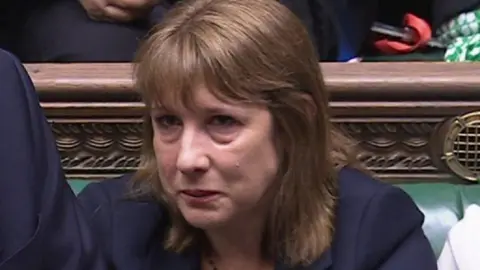 Rachel Reeves sat in Parliament, with tears visibly coming down her cheeks. She has short brown hair with a fringe and is wearing a suit.