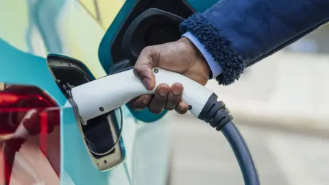 A person putting an electric vehicle charger into the side of a light blue car.