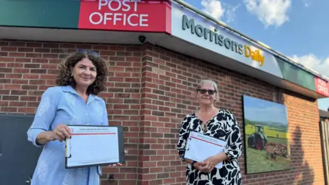 Diana Johnson MP Two women stand together outside the Morrisons Daily store, holding on paper petitions on clipboards. The woman on the left has curly, dark hair and sunglasses on her head, and wears a light blue dress. The woman on the right has blonde hair tied back, sunglasses on her face, and weas a black and white patterned dress.