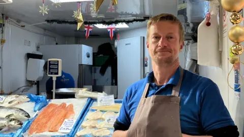 A man stands in front of fish market stall decorated with Christmas ornaments and garlands, featuring a display of fresh seafood on ice. There are British flags hanging above the counter. He has his hands tucked behind a brown apron and is wearing a blue polo shirt.
