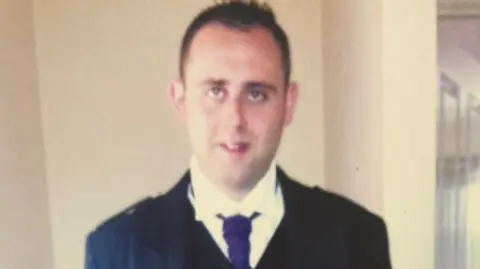 Head and shoulders shot of a pale looking man with short dark hair. He is smiling and looking at the camera. He is wearing a dark kilt jacket, a white shirt and a purple neck tie. The man appears to be standing in a domestic setting against a white wall.