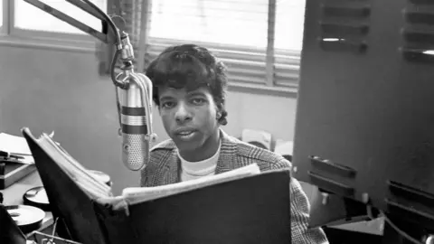 Getty Images A young Sly Stone wears a tweed jacket and sits in front of an old-fashioned microphone with a folder in front of him. He looks into the camera.