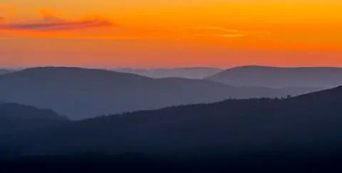 Getty Images A deep red sunset with forest covered hills beneath. There are a very few wispy clouds in the sky.