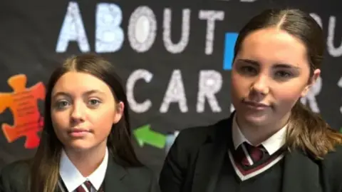 Two teenage girls with brown hair, wearing black school uniforms