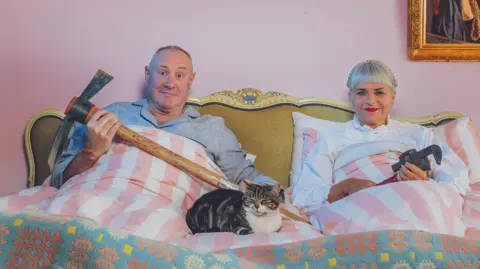 Keith and Marj are sat up in a bed with pink and white striped bedding. There is a cat sat between them. Both of them are holding building tools. The wall behind them is pink and has a hole in it exposing wooden panels. On the same wall is a gold mirror.