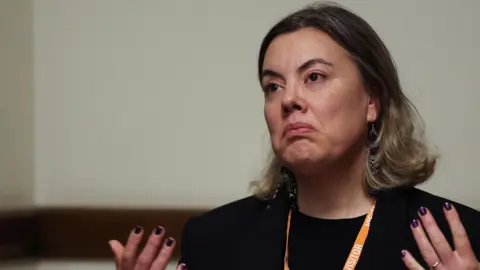 Reuters Greenland's Minister for Business, Mineral Resources, Energy, Justice and Gender Equality Naaja Nathanielsen speaks at a press conference, wearing a black top and an orange lanyard around her neck.