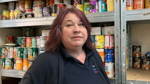 A woman with dark purple hair, a black polo shirt and black cardigan looks at the camera with a neutral expression. Behind her is a shelf full of tinned foods.