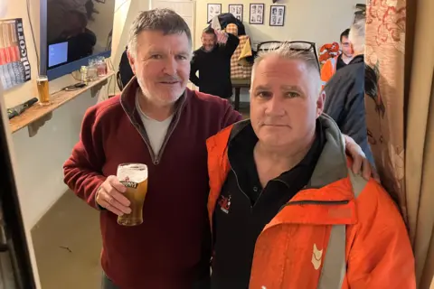 Two men stand inside a flooded pub, posing for the camera. The man on the left has a pint of beer in his right hand and his left arm is around the man to his right. Both men have short grey hair. Brown water can be seen in the background, about ankle height. There is a television in the background and three other men.