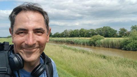 A selfie of Karl Lansley, who is smiling at the camera, taken beside a river running through a field