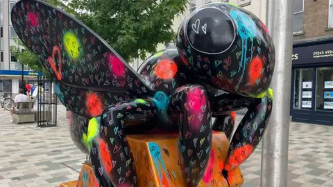 Steve Huntley/BBC A bee sculpture painted in black with small colourful hearts drawn on it and spray-painted patches in luminous colours. It is standing in a pedestrianised street lined by shops.