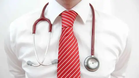 A close up of a man's chest. He is wearing a white shirt and red tie and has a red and silver stethoscope hanging around his neck.