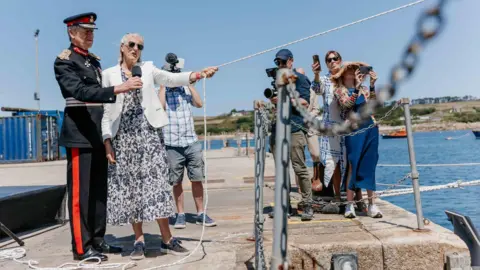 A man in uniform of black and red with a hat holds a microphone in front of a woman holding the vessel's rope while to their right are bystanders holding phones and cameras, with the sea in the background. 