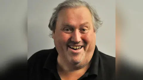 Stoke Amateur Theatre Society Paul Brammer, a man with grey hair is smiling. He is wearing a black polo shirt, and is stood against a pale coloured backdrop.