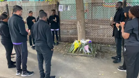 A group of people dressed in black stand in a circle around a tree, at the foot of which they have laid floral tributes.