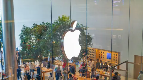 Getty Images The Apple logo is pictured on the glass wall of an Apple store with people shopping below