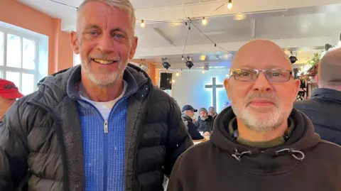 Mark Butcher (left) with grey hair and beard wearing a white t-shirt, blue jumper with a zip and black puffer jacket and Mark Clarey with a grey beard wearing silver glasses. He is wearing a green fleece underneath a brown hoodie. They are both smiling. They are standing in Pizza Grazia with regulars sitting at tables eating pizza behind them.