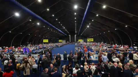 PA Crowds of people stand on a blue carpet inside the Titanic Exhibition Centre where votes are counted for local elections. There are yellow signs for different constituencies around Belfast. 