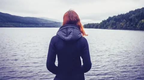 Getty Images Young woman with her back to the camera looking out over a misty Scottish loch - generic. She has red hair and is wearing a blue jacket with a hood.